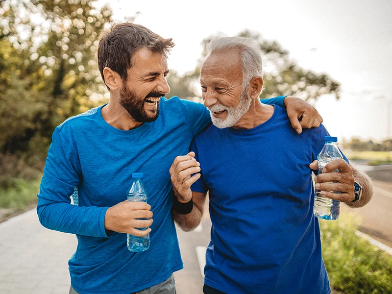 Two men, one younger and one older, walk together outdoors with their arms around each other, smiling and holding water bottles on a sunny path. (photo)