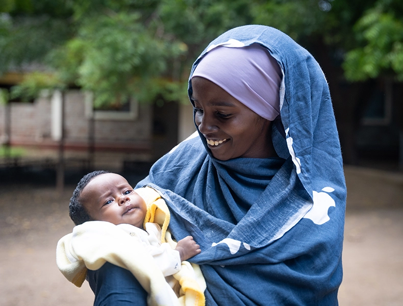 A woman smiling while holding a baby outdoors. (photo)