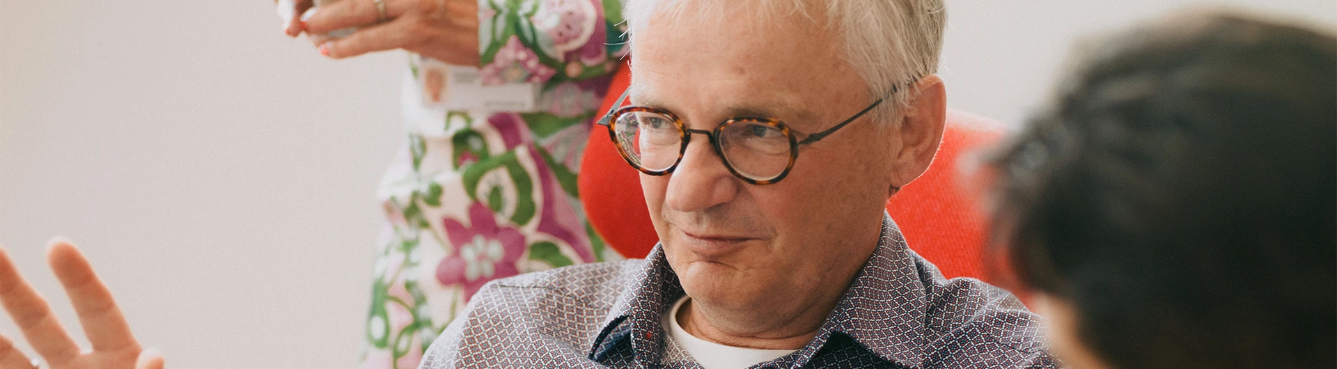 An older man wearing glasses gestures while speaking to someone in a bright indoor setting. (photo)