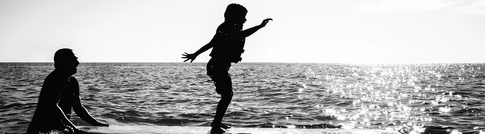 A silhouetted child jumps into the sea while an adult watches nearby, with sunlight shimmering across the water. (photo)