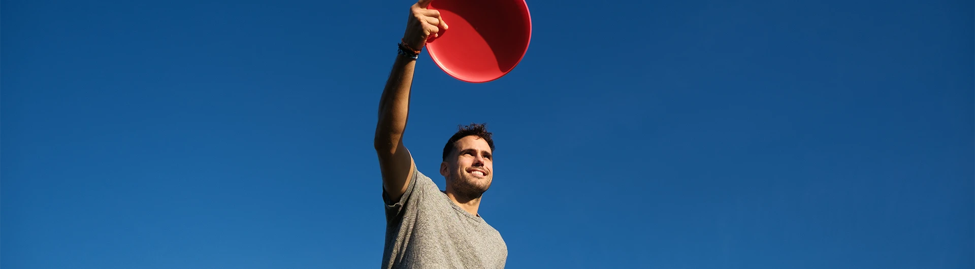 A man smiles while holding a red flying disc up against a clear blue sky. (photo)