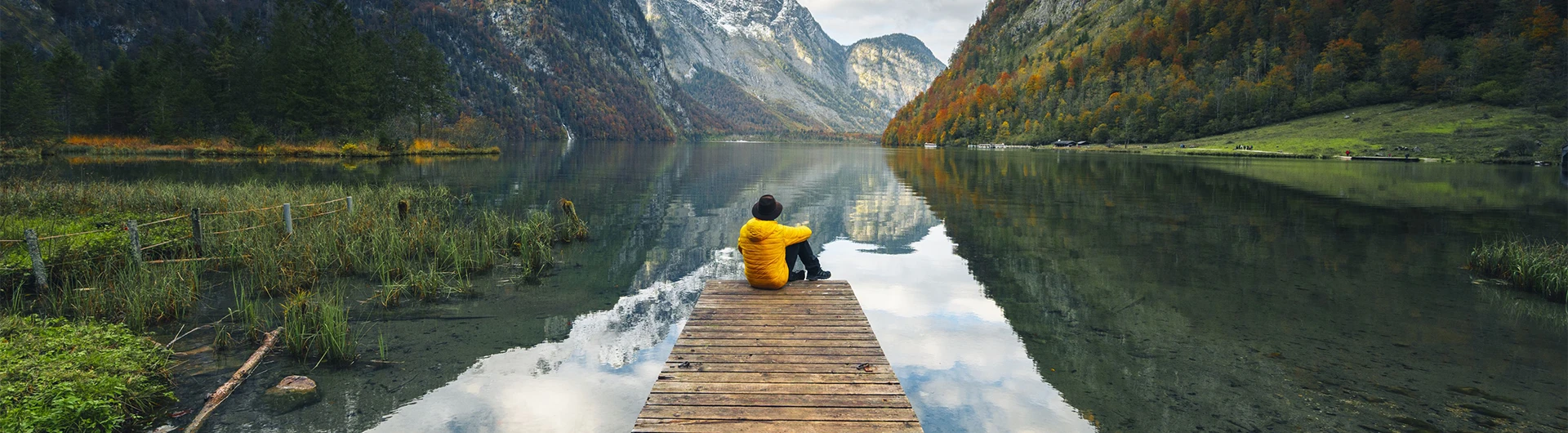 A person in a yellow jacket sits on a wooden dock overlooking a calm mountain lake surrounded by autumn trees. (photo)