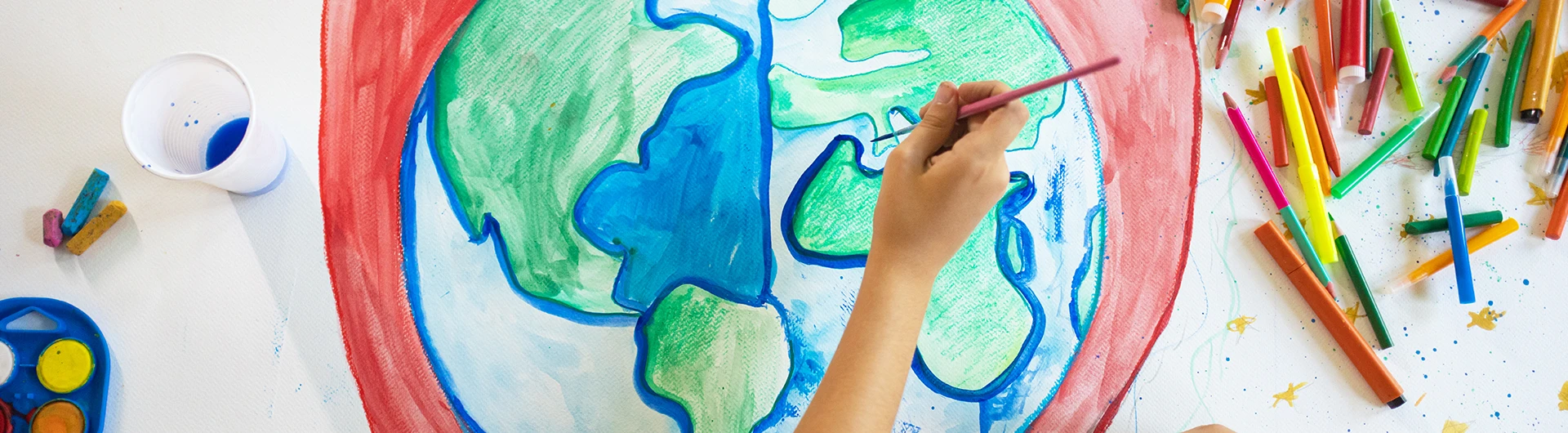 A child’s hand paints a colorful picture of the Earth, surrounded by crayons and art supplies on a table. (photo)