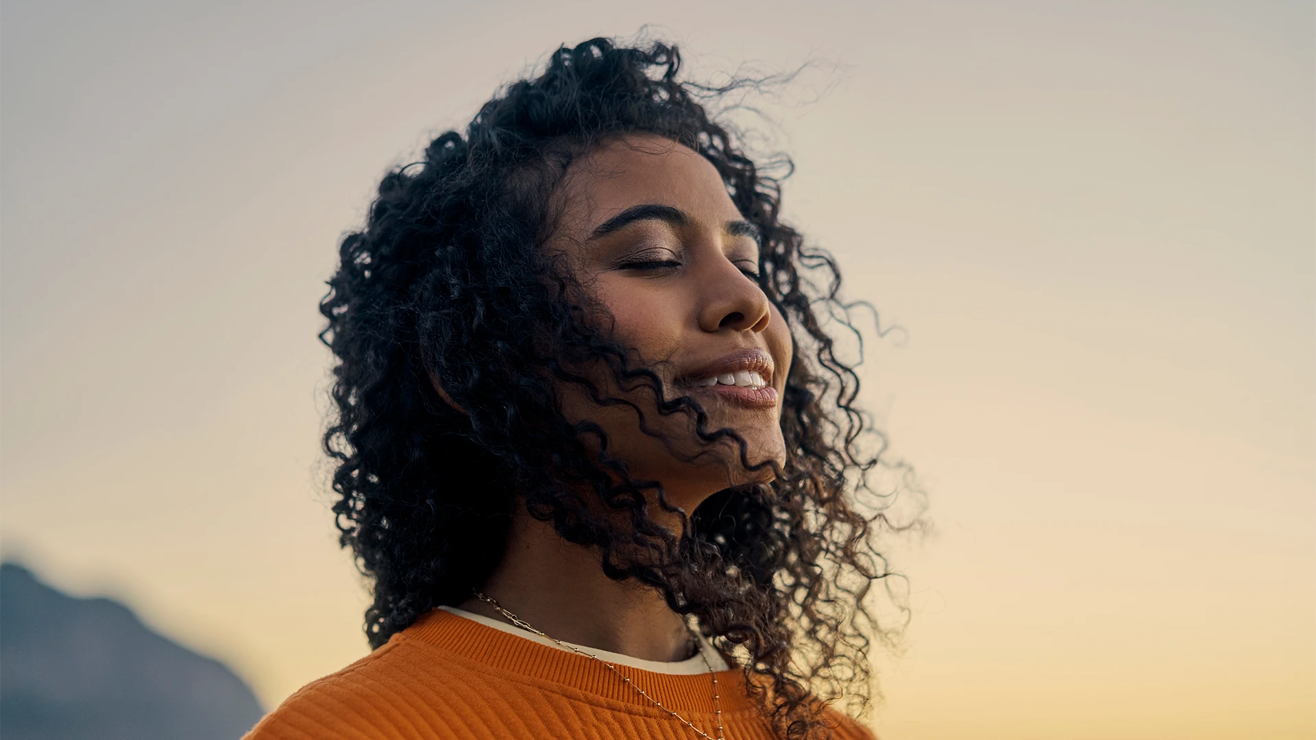 A woman with curly hair smiles softly with her eyes closed outdoors at sunset. (photo)