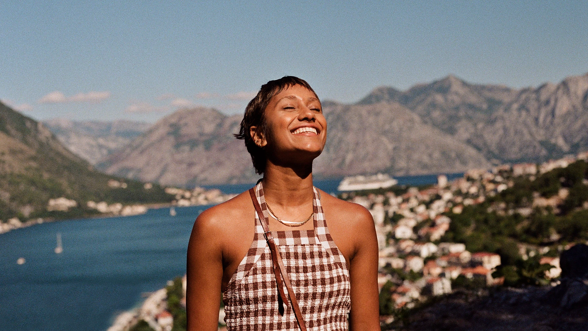 A woman smiles with her eyes closed while standing above a coastal town, with mountains and blue water in the background. (photo)