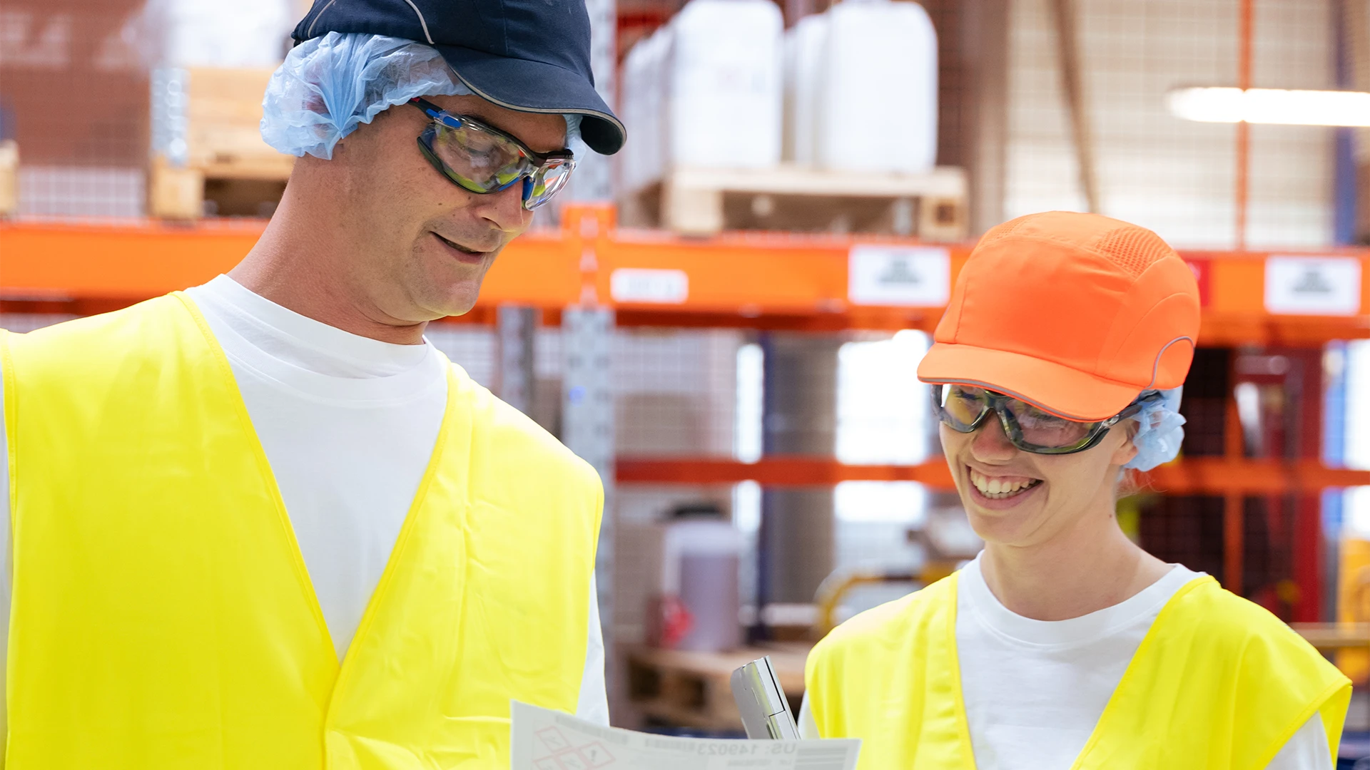 Two warehouse employees wearing safety vests, caps, hair nets, and protective glasses review a document together, smiling as they stand in a brightly lit industrial setting. (photo)