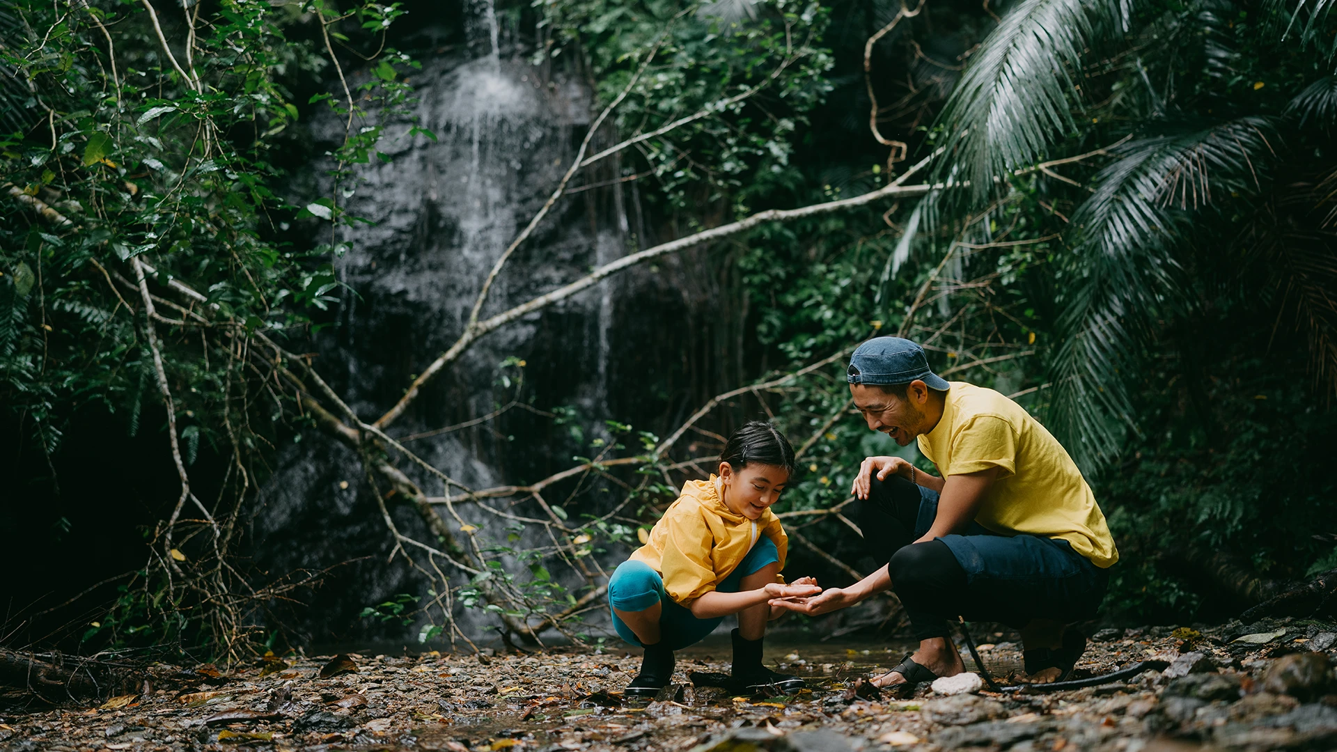A parent and child crouch by a shallow stream in a lush forest, smiling as they explore the water together near a small waterfall. (photo)