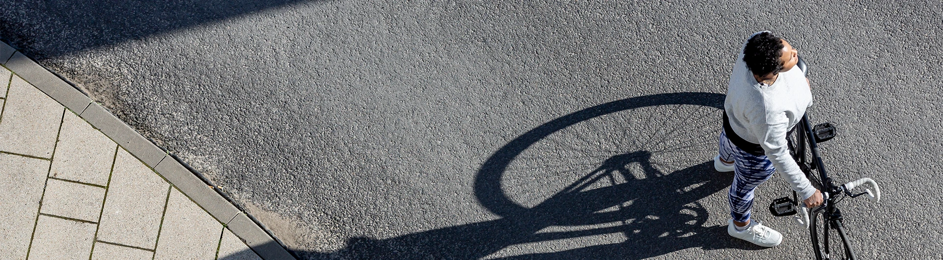 A person walks their bicycle along a sunlit street, seen from above, with a long shadow stretching across the pavement. (photo)