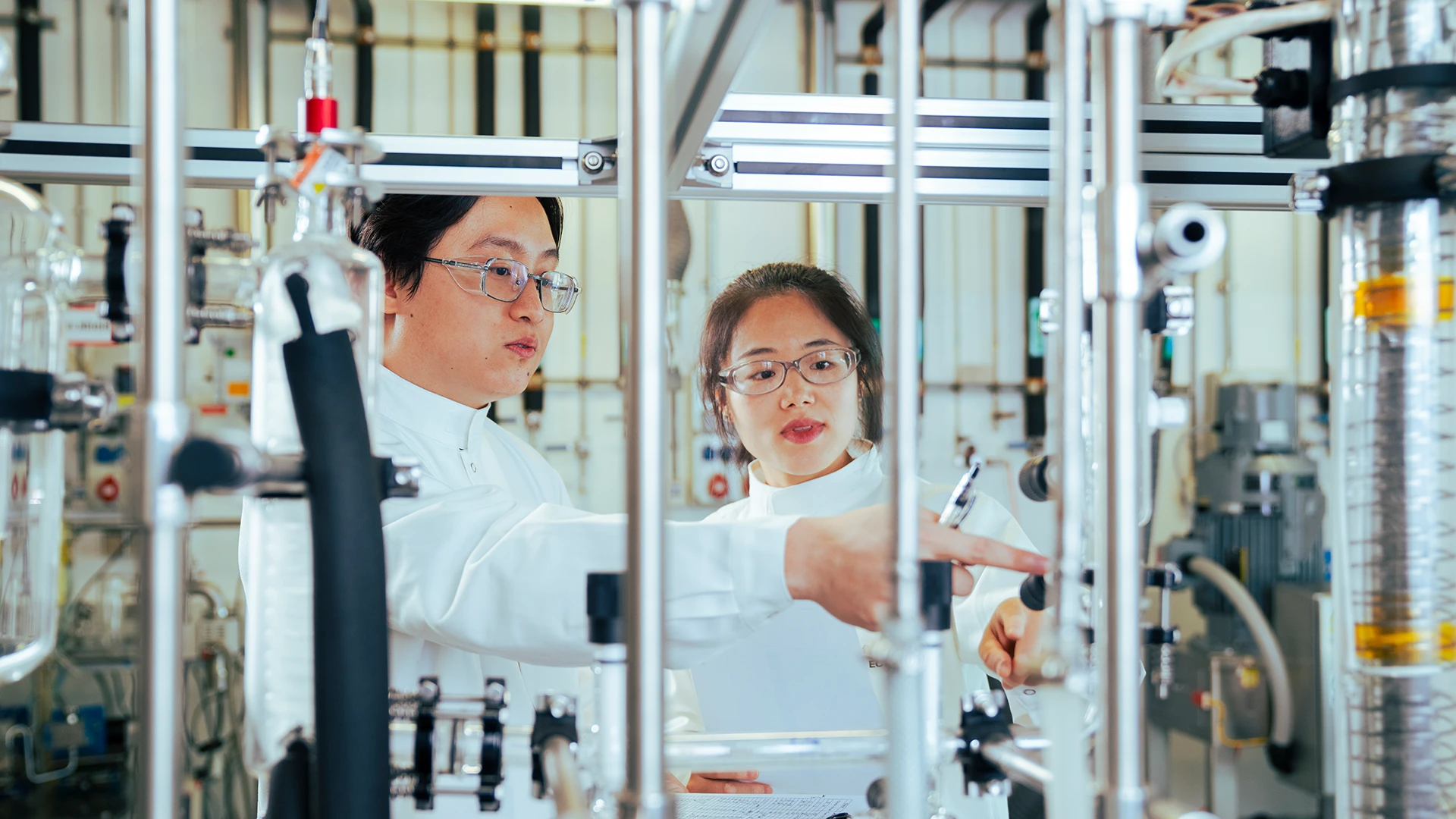 Two scientists in protective lab wear discuss and point at equipment within a complex laboratory setup. (photo)