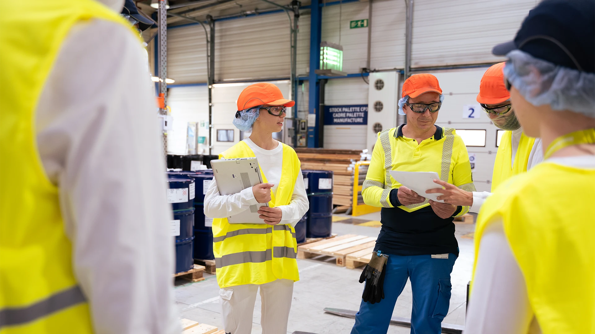 A group of factory workers in safety gear review documents together on a production floor. (photo)