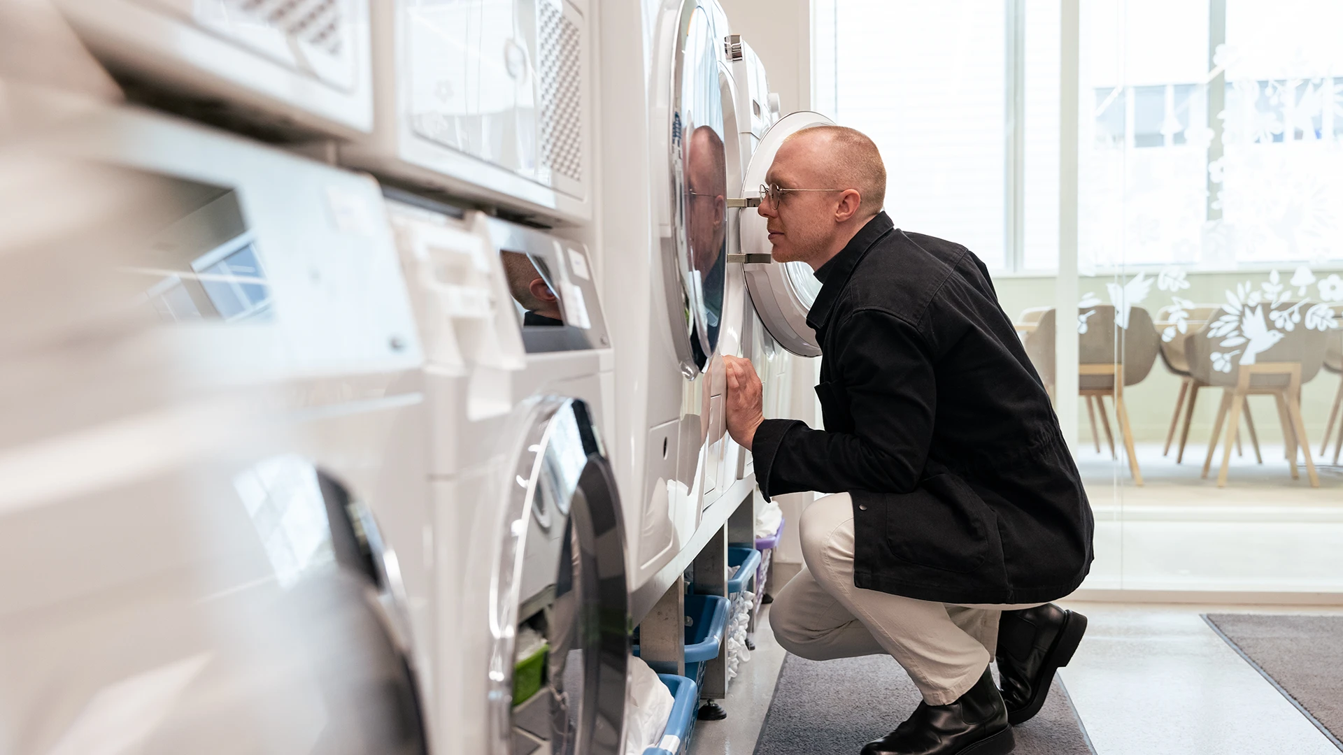 A man crouches in a bright showroom, closely inspecting a washing machine behind a glass door. (photo)