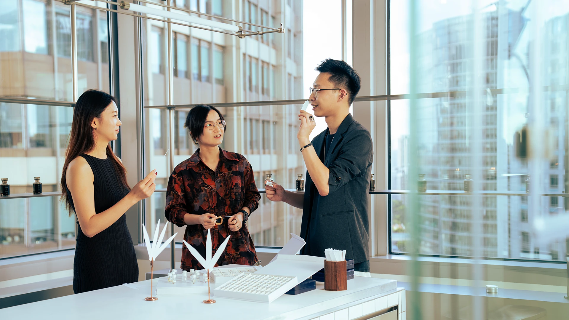 Three colleagues evaluate and discuss fragrance samples in a bright studio with large windows overlooking the city. (photo)