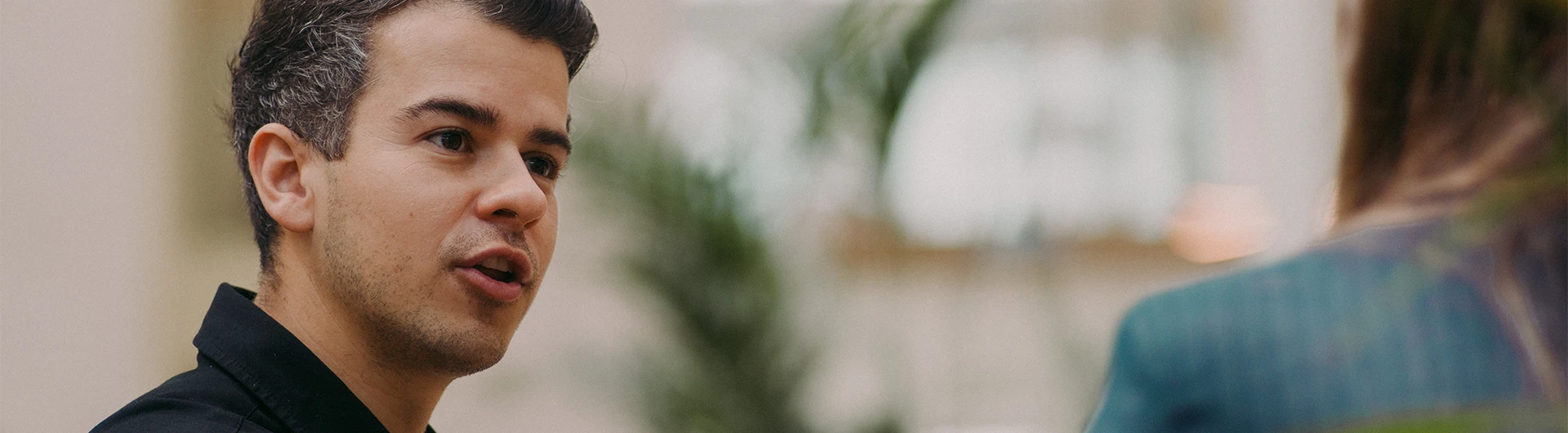 A man speaks to another person in a softly lit indoor setting with plants in the background. (photo)