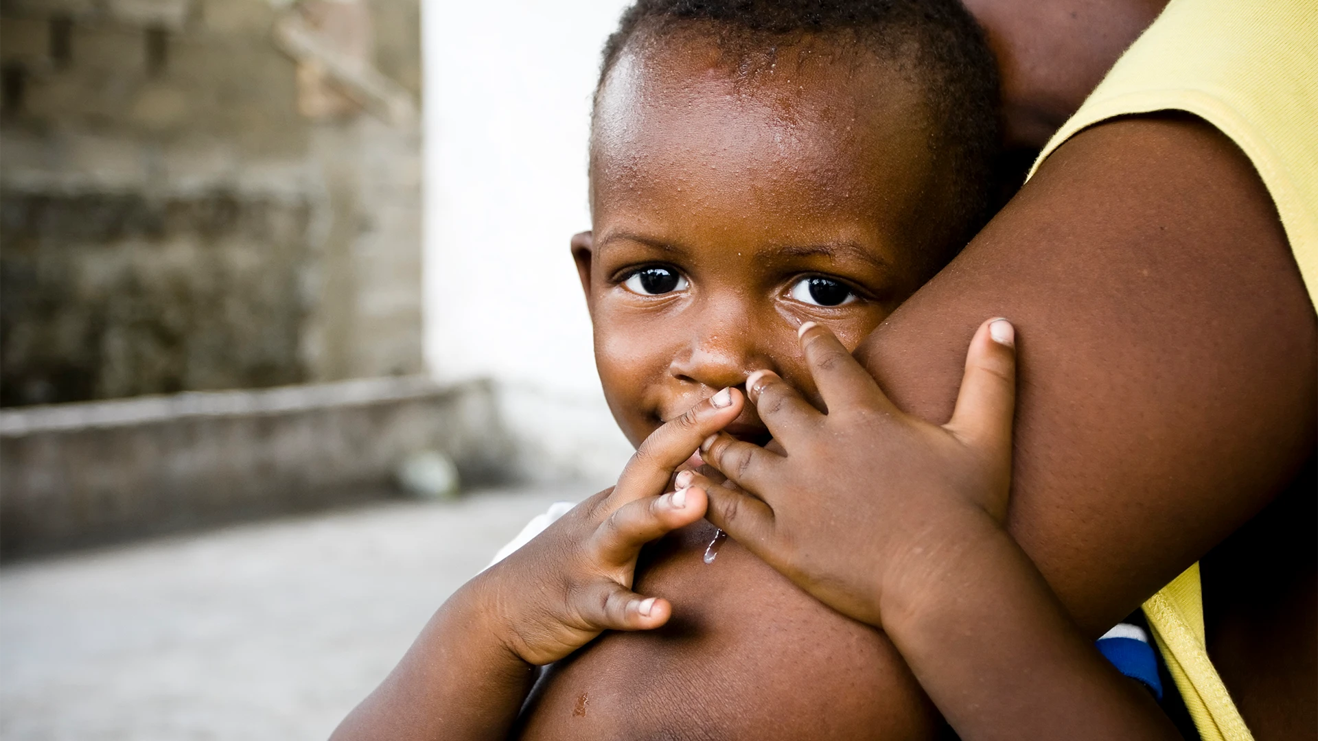 A young child peeks shyly toward the camera while being held close by an adult, resting against their arm in a quiet, intimate moment. (photo)