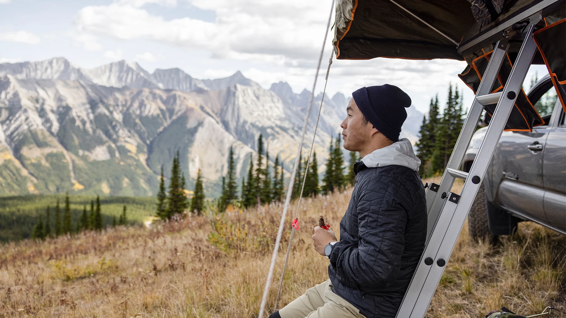 A man pauses beside a rooftop tent, enjoying a quiet moment and a snack while gazing out over a vast mountain landscape. (photo)