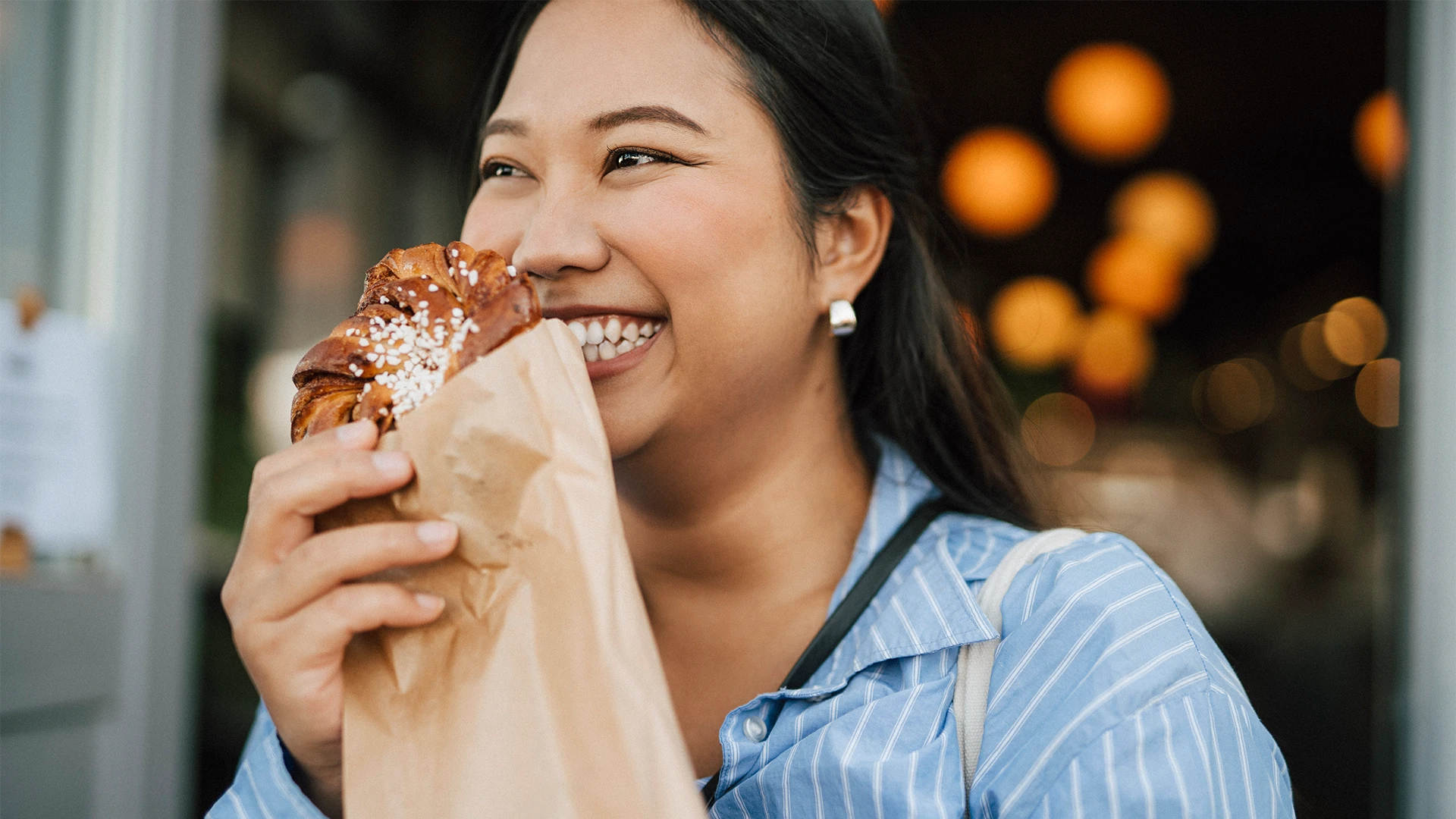 A woman smiles as she takes a bite of a freshly baked pastry held in a paper bag, standing outside a café with warm, softly glowing lights in the background. (photo)