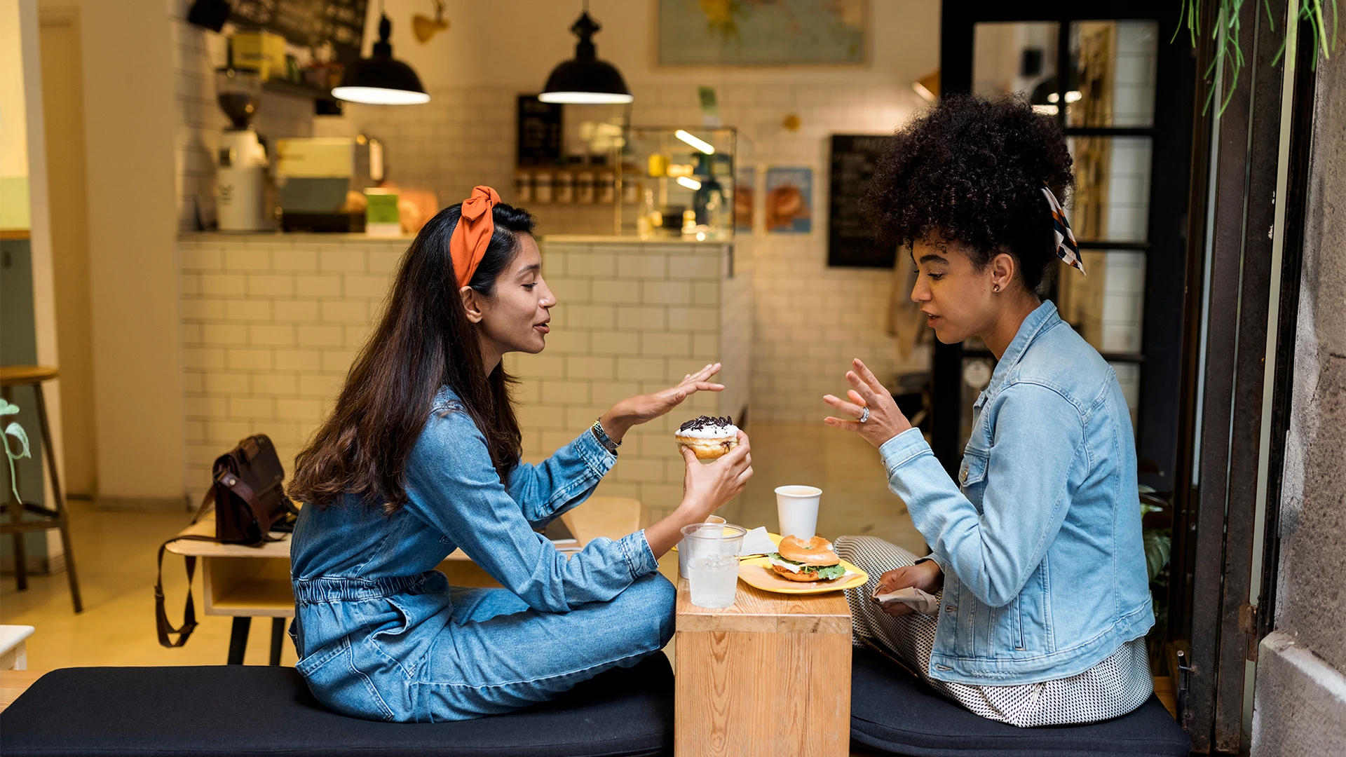 Two people sit at a café table, chatting and sharing food and drinks in a cozy, casual setting. (photo)