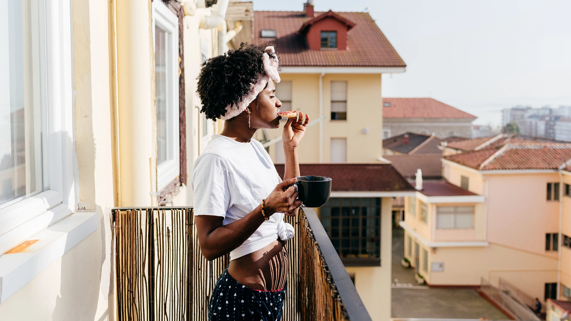A woman enjoys a quiet morning on a balcony, sipping coffee and eating breakfast while overlooking city rooftops. (photo)