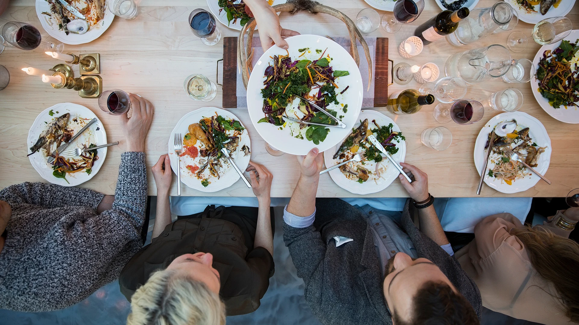 People share a meal around a table as a fresh salad is passed between plates, viewed from above. (photo)