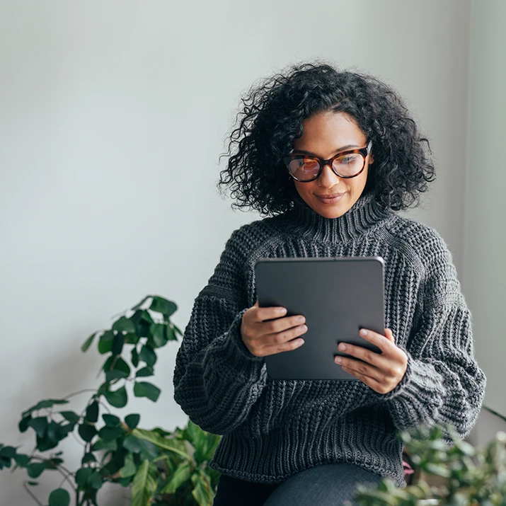 A woman with glasses reads content on a tablet while standing indoors near green plants. (photo)