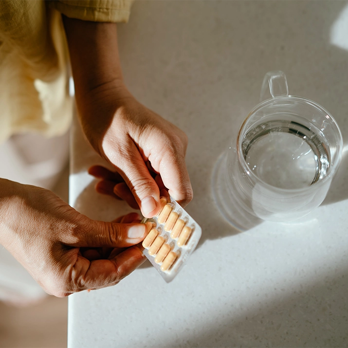 Hands holding a blister pack of capsules next to a glass of water. (photo)